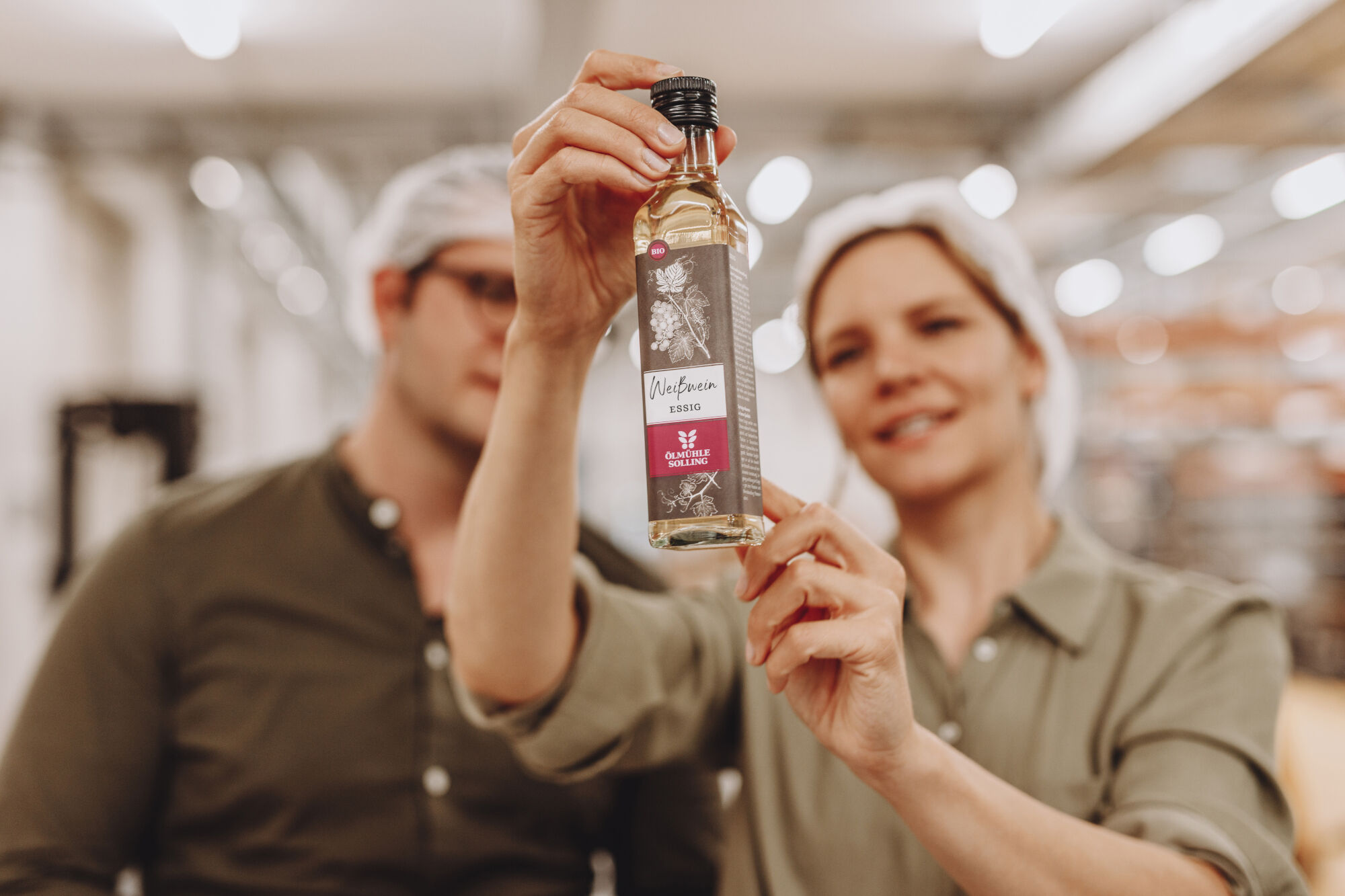Person hält eine kleine Glasflasche mit Etikett 'Ölmühle Solling' und rotem Logo in einer Produktionshalle